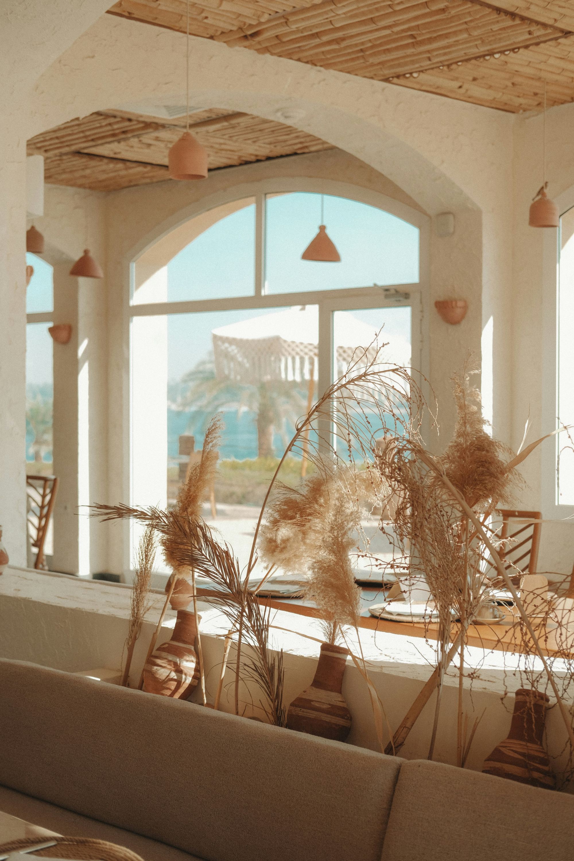 An Ibiza villa interior with whitewashed walls, a bamboo-and-reed ceiling, terracotta pendant lights, and an arched window opening onto a palm tree and the Mediterranean sea.