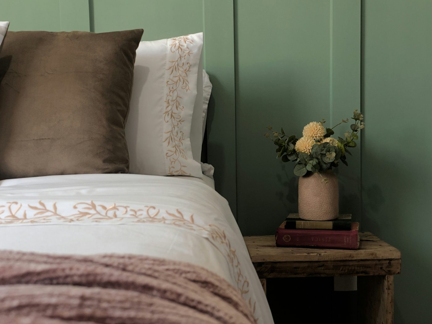 A guest bedroom with sage panelled walls, embroidered white linens, and a small vase of dahlias on a wooden bedside table.